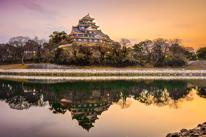 A Japanese castle with tiered roofs is reflected on a calm river at sunset, surrounded by trees and stone walls, creating an enchanting scene reminiscent of ancient citadels.