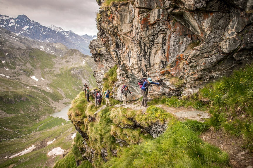 Hikers traverse a narrow, rocky mountain path with a steep drop-off. Snow-capped peaks and a green valley are visible in the background under a cloudy sky, as they follow the famous Haute Route between France and Switzerland.