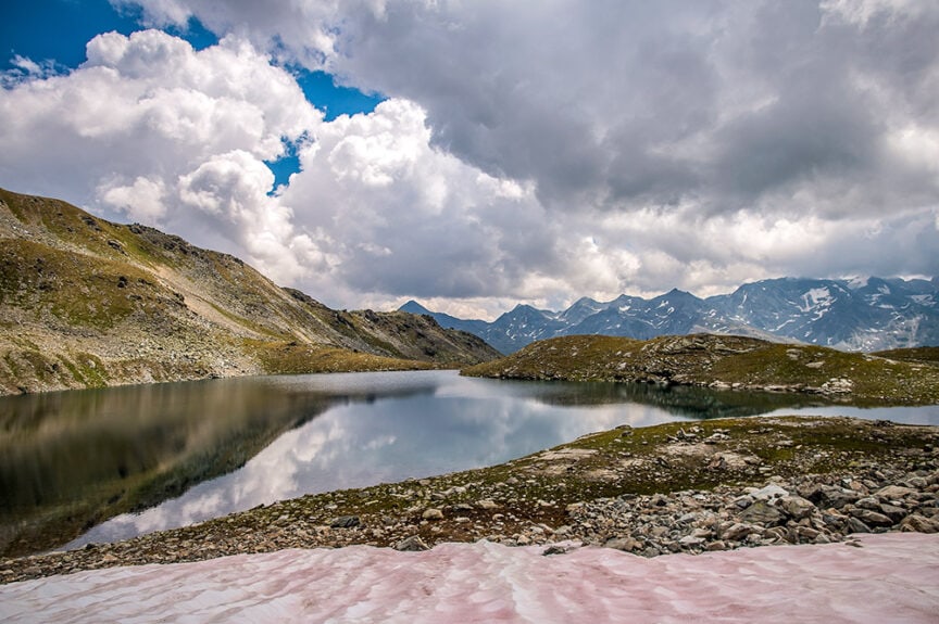 A serene mountain lake surrounded by rocky terrain with snow patches in the foreground, under a partly cloudy sky with mountains in the distance, reminiscent of the Haute Route between Switzerland and France.