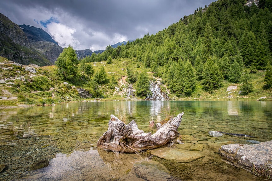 A serene mountain landscape along the Haute Route with a clear lake in the foreground, a fallen tree in the water, and a waterfall surrounded by green trees and rocky mountains in the background under a partly cloudy sky.