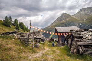 Rustic wooden huts in a mountainous area, with Swiss flag bunting strung between poles. A cloudy sky and rocky peaks are in the background, capturing the essence of the Haute Route between France and Switzerland.