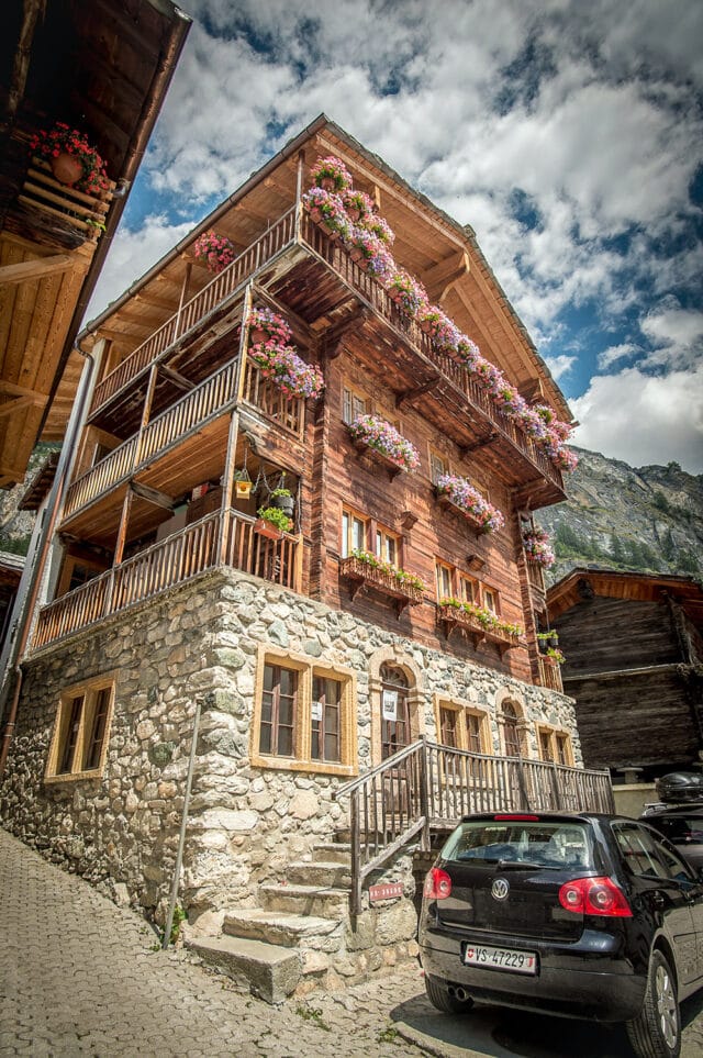 A three-story wooden house with a stone foundation, adorned with flower pots on balconies, reminiscent of a quaint Swiss chalet. A black car is parked in front. The background features mountains and a partly cloudy sky, evoking the picturesque landscapes along the Haute Route through Switzerland and France.