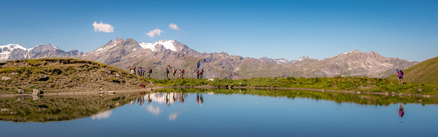 A group of hikers walks along a grassy hill overlooking a calm lake, with snow-capped mountains in the background under a clear blue sky, reminiscent of the scenic trails found on the Haute Route between France and Switzerland.