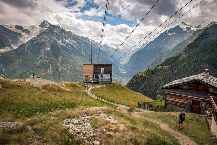 A small mountain cable car station in France, surrounded by lush greenery, tall mountains, and cloudy skies, with a few people walking along a path leading to the station.