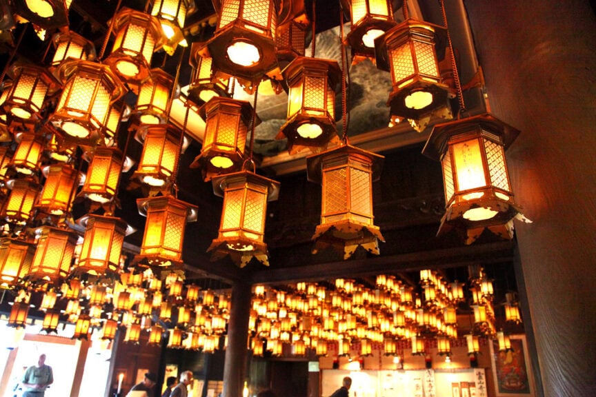 An indoor space with numerous hanging lanterns creates a warm, ambient light akin to a serene evening in Japan. Visitors in the background, reminiscent of modern American Pilgrims, engage with the captivating environment.