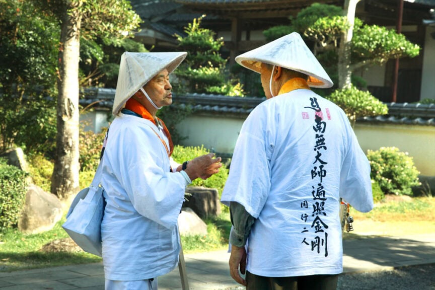 Two people wearing traditional conical hats and white robes with Japanese writing stand facing each other outdoors, engaged in conversation, embodying the cultural exchange between Japan and an American pilgrim.