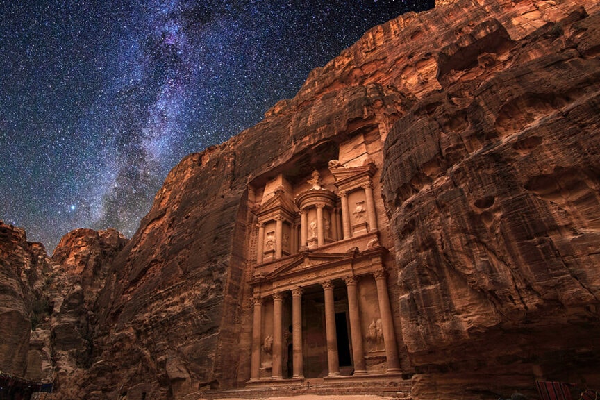 The Treasury at Petra, Jordan, illuminated at night under a starry sky with the Milky Way visible above the rock-cut architecture, is a stunning example of urban antiquity.