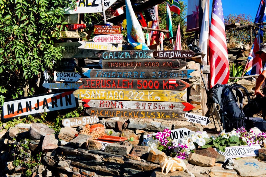 A colorful assortment of directional signposts, each proclaiming Buen Camino, points to various cities and distances like Santiago, Jerusalem, and Rome, all surrounded by rocks and flags.