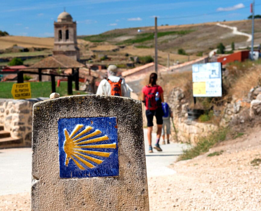 A stone marker with a yellow scallop shell symbol identifies the Camino de Santiago route. Pilgrims, often greeting each other with "Buen Camino," walk along the path towards a village with a bell tower.