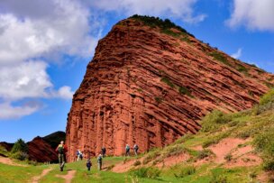A group of hikers embark on an adventure, walking on a path towards a large, red sandstone rock formation under a partly cloudy blue sky in the breathtaking landscapes of Kyrgyzstan.