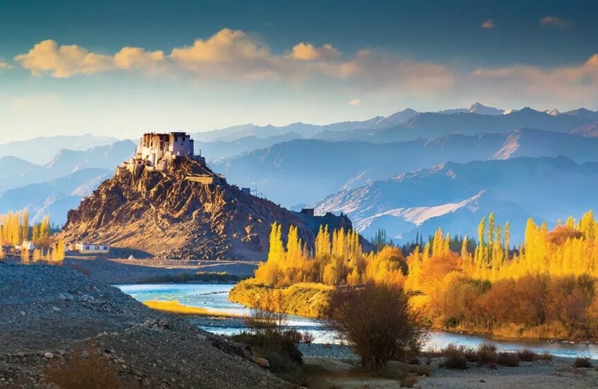 A fortress atop a hill overlooks a river with autumn foliage, set against a backdrop of mountains under a blue sky—a perfect destination for adventurous hiking trips that promise breathtaking views and exciting trails.