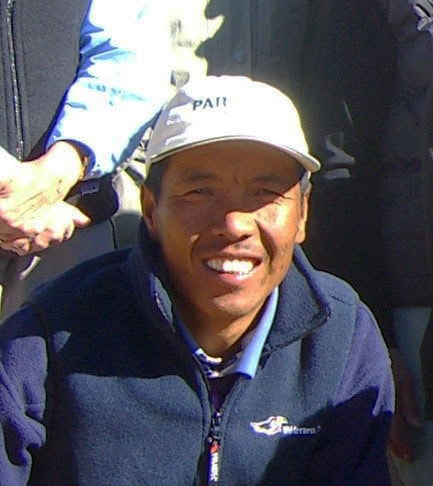 A man wearing a light-colored cap and a dark jacket, smiling, is captured in a close-up outdoor photograph that would be perfect for a family photobook from Nepal.