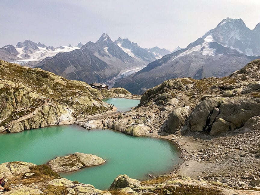A mountain landscape with turquoise lakes, rocky terrain, and snow-capped peaks under a partly cloudy sky. A few people are visible hiking on the paths and near the water, perhaps as part of their Tour du Mont Blanc adventure.