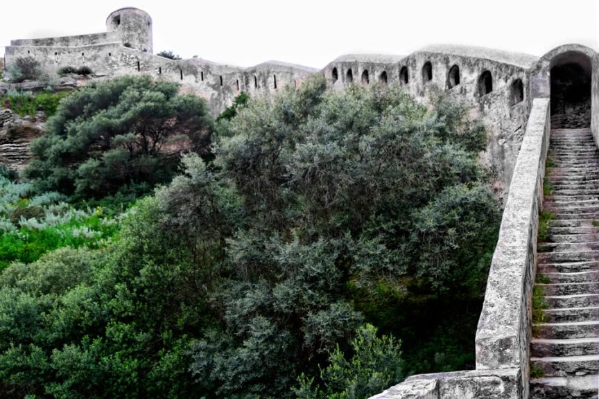 An ancient stone fortress with battlements and an arched passage, surrounded by dense, green foliage and trees—a scene worth a thousand pictures. Rocky steps lead up alongside the majestic structure.