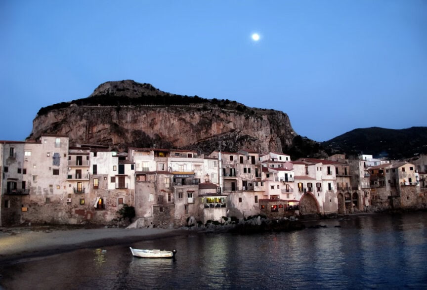 A coastal village at dusk, illuminated by building lights and a full moon, looks like a scene from a thousand words. A small boat on calm water graces the foreground while rocky hills rise in the background, painting a picturesque moment that feels timeless.
