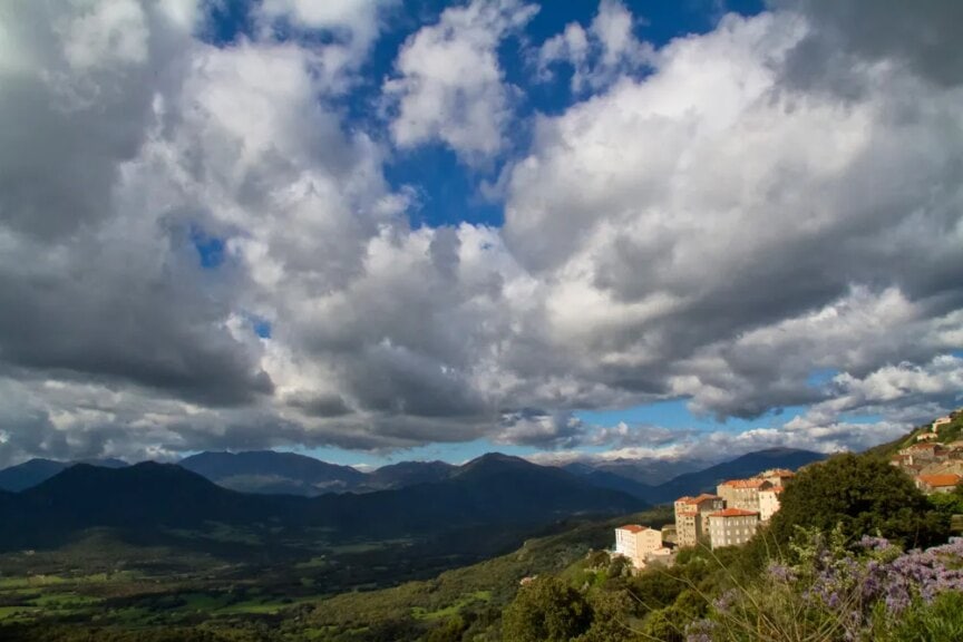 A hillside town under a partly cloudy sky, with a vast green valley and mountain range in the background, is worth a thousand words.