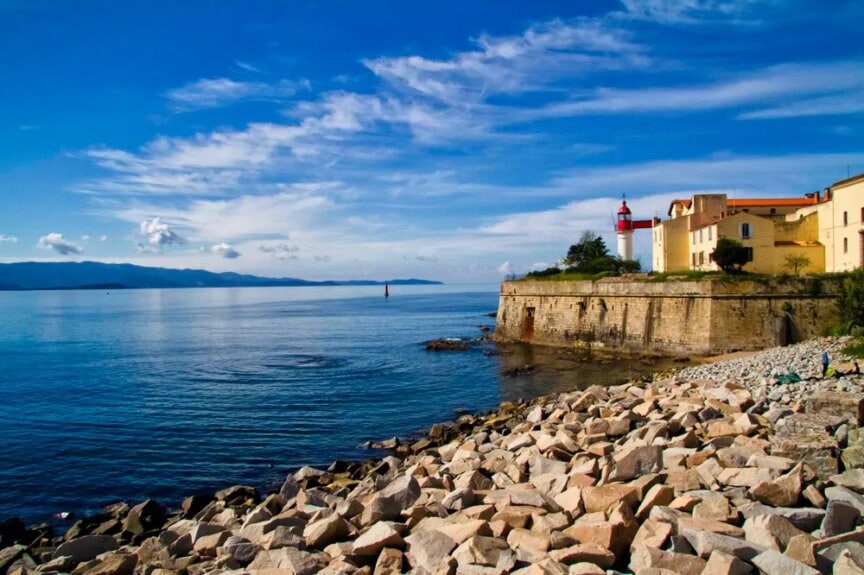 Rocky shoreline with calm sea, a red lighthouse, and yellow buildings on a raised platform under a blue sky with scattered clouds—pictures that truly speak a thousand words.