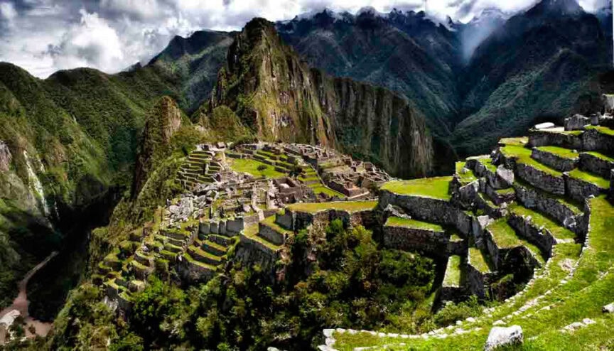 A panoramic view of Machu Picchu, featuring ancient stone structures and terraces set against a backdrop of lush green mountains and clouds, is worth a thousand words.