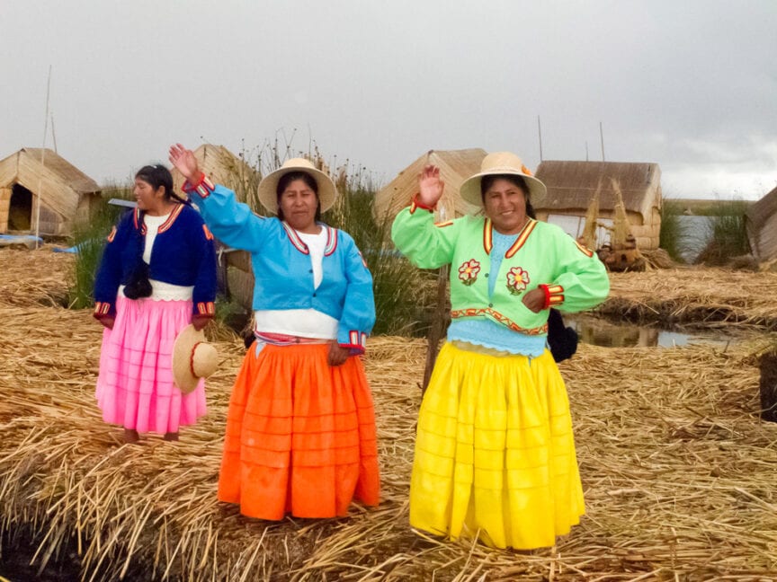 Three women in brightly colored traditional outfits stand on a straw-covered surface, waving. Small straw huts form the backdrop of this picturesque scene. It's said that pictures speak a thousand words; this vibrant moment certainly tells its own story.