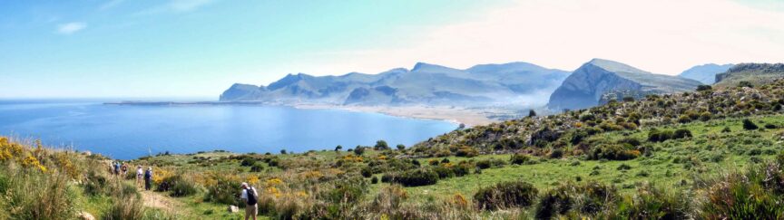 A group of people hike along a verdant coastal path overlooking a calm blue sea and mountain range on a clear, sunny day, each step capturing moments worth a thousand words.
