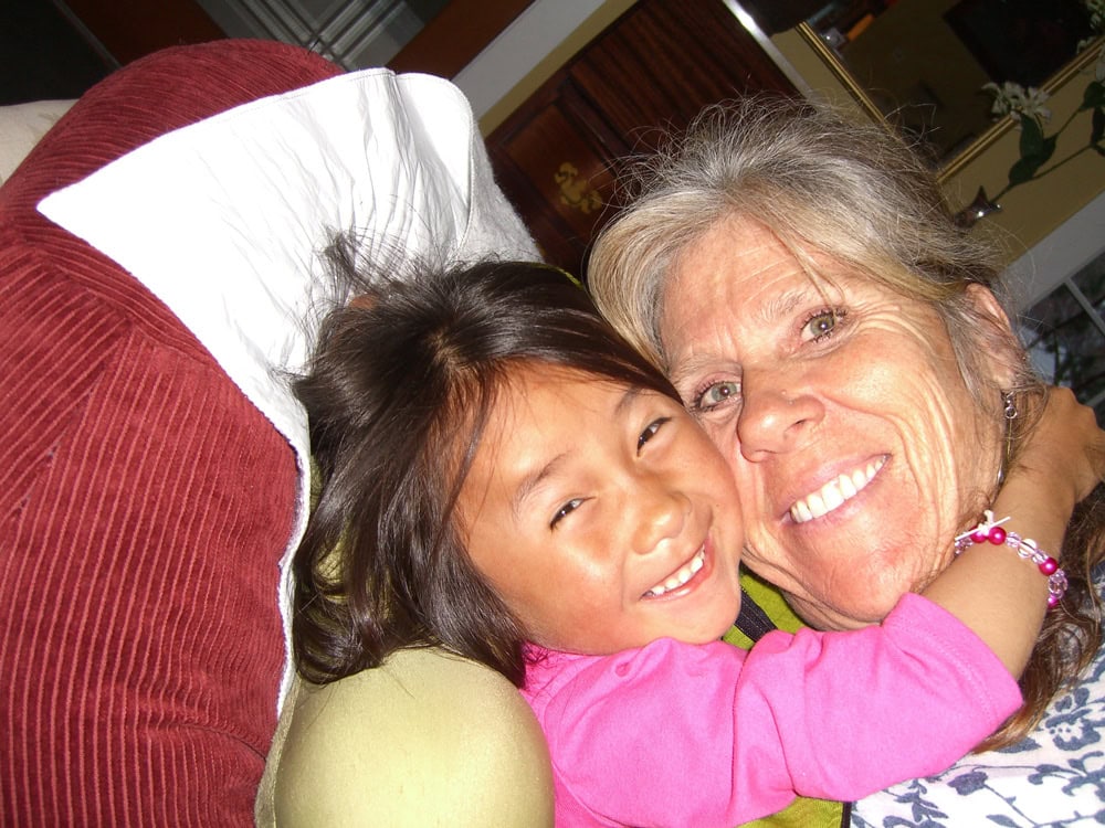 A child in a pink shirt and an older woman are smiling and hugging each other, capturing a precious family moment perfect for their photobook while sitting indoors.