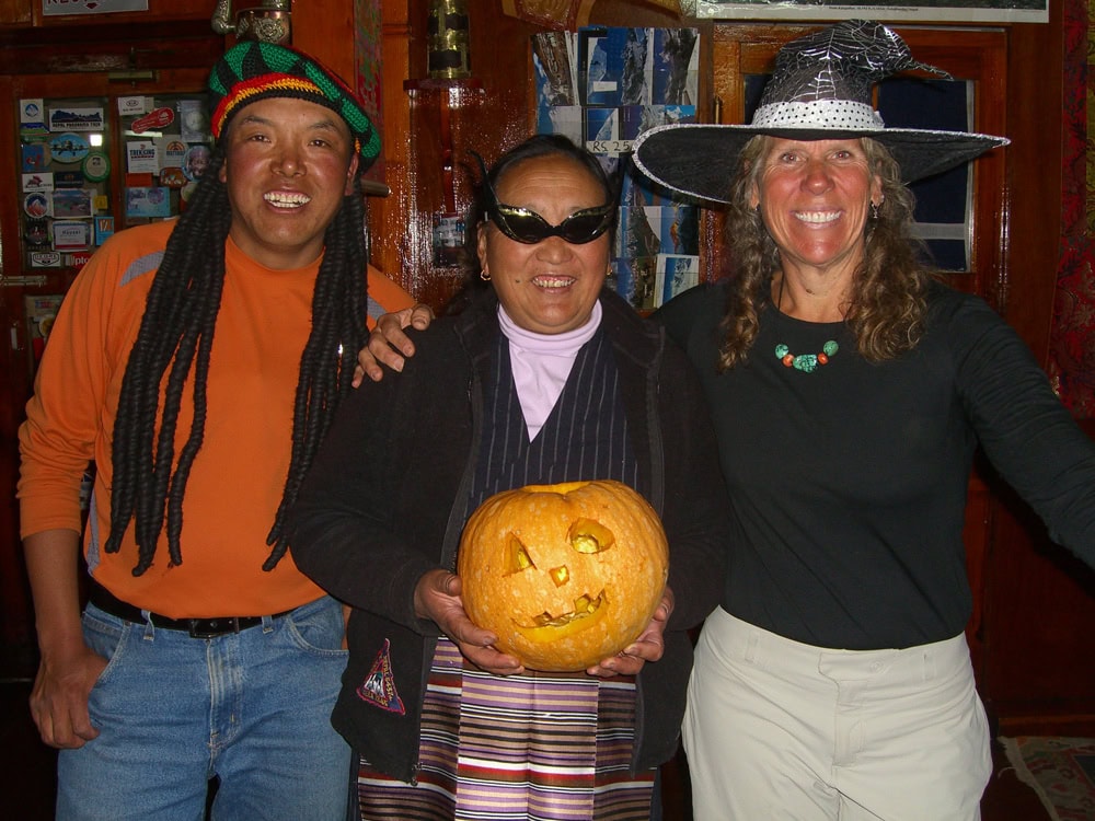Three people in costume, including a man wearing a multicolored hat with dreadlocks, a woman with sunglasses holding a carved pumpkin, and another woman in a witch's hat, smiling inside a room. This lively scene could be straight out of a cherished family photobook.