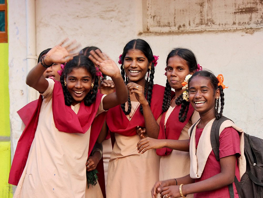 A group of schoolgirls in uniforms, with red scarves and flower-adorned braids, smile and wave at the camera while standing against a light-colored wall. This cheerful portrait captures the vibrant essence of South India.