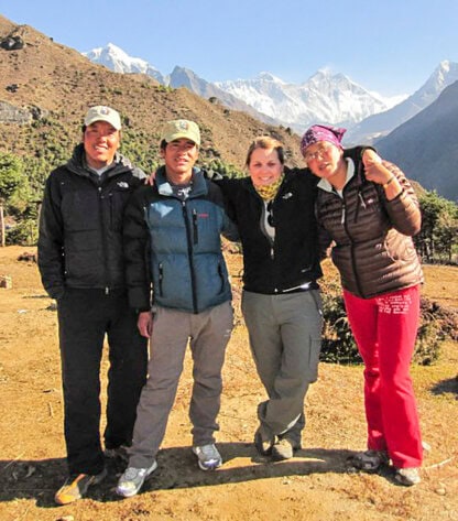 Four people in outdoor gear pose together on a mountain trail in Nepal, with snowy peaks in the background, capturing the spirit of exploration.