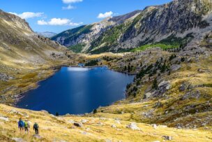 Three hikers walk through a grassy valley towards a large lake nestled among rocky mountains under a blue sky, enjoying their hiking adventure.