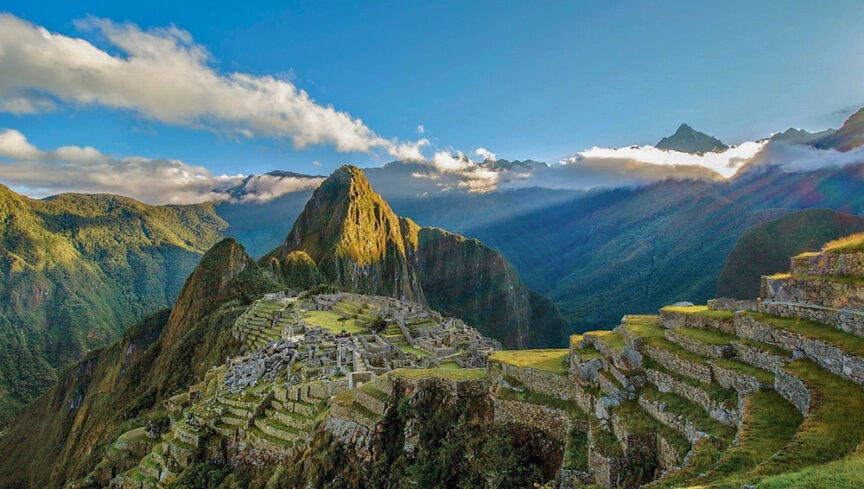 A view of Machu Picchu with its stone structures and terraces surrounded by lush green mountains under a cloudy blue sky—a must-see on any 9-day travel adventure.