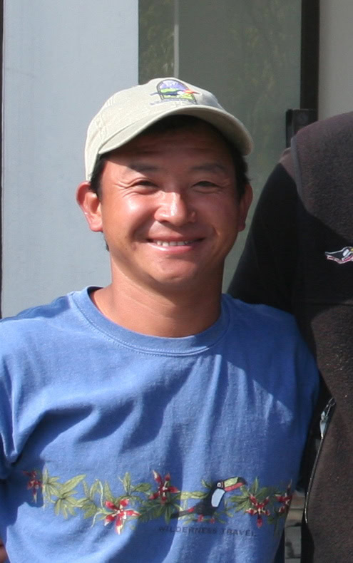 A man wearing a blue T-shirt with a floral design and a light-colored cap is smiling at the camera, capturing a joyful moment for the family photobook during their trip to Nepal.