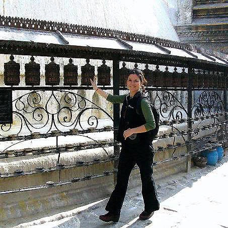 A person stands beside a row of prayer wheels in Nepal, touching one wheel with their hand, in front of a decorative metal fence at a religious site—a serene moment captured perfectly for the family photobook.