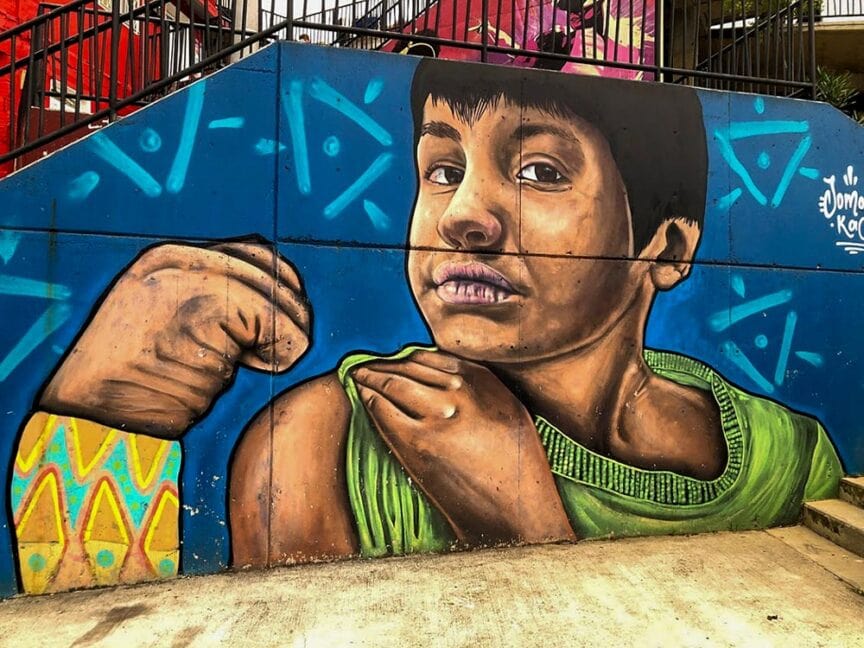 A mural on a building wall in Colombia depicts a child with short dark hair, looking sideways, and revealing an arm with a rolled-up sleeve, surrounded by vibrant blue and green geometric patterns. This artwork offers amazing experiences for all who encounter its captivating design.