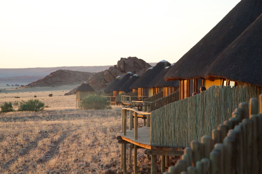 A row of thatched-roof huts with wooden fences on an arid, grassy landscape at sunset, set against the rocky hills of Namibia's wonderland adventure.
