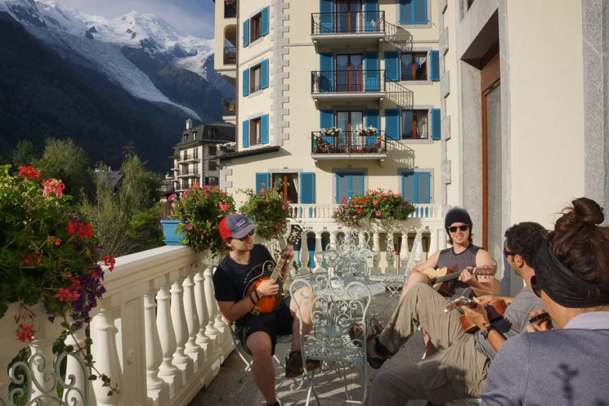 Four people are seated on a balcony with decorative railings, playing string instruments. In the background, there are buildings, green trees, and mountains with Mt. Blanc's glacier looming under a partly cloudy sky.