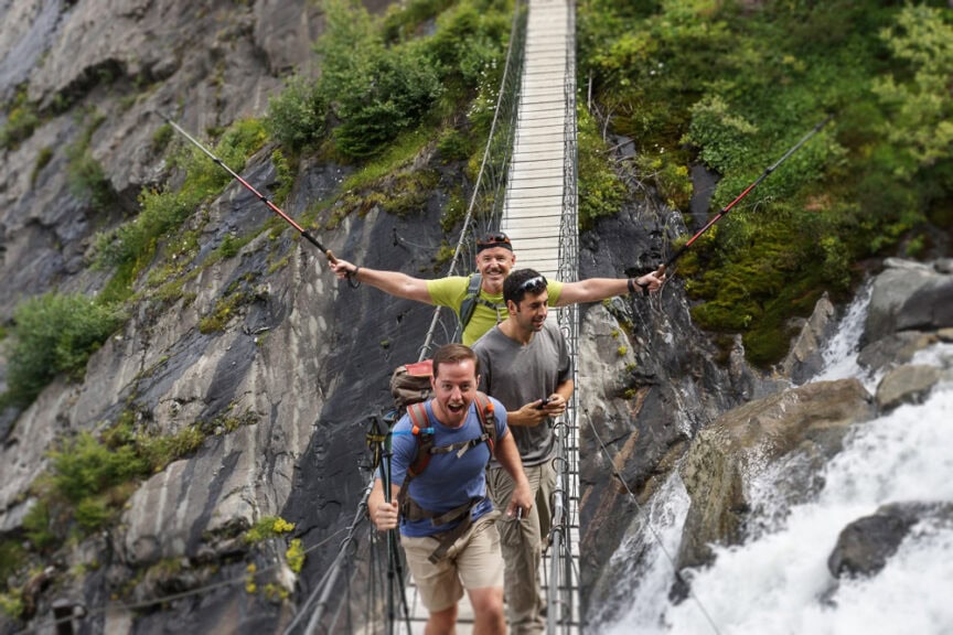 Three people circumnavigating a suspension bridge over a waterfall, smiling and holding hiking poles. The background features rocky terrain and greenery, reminiscent of an adventure near Mt. Blanc.