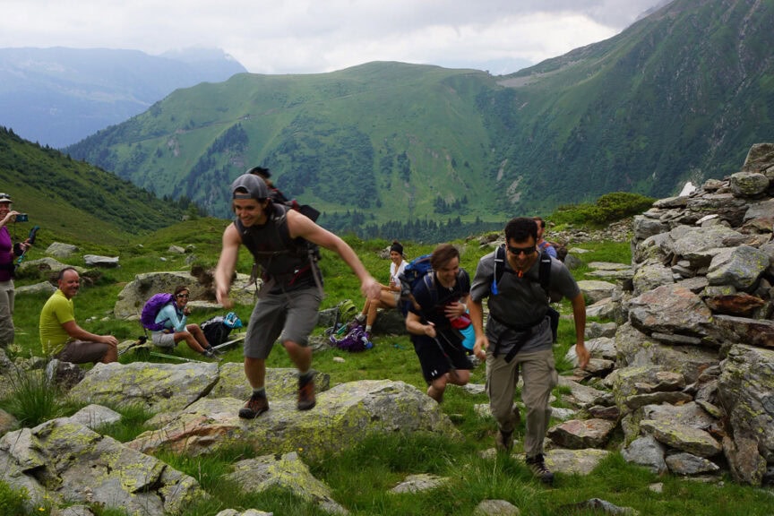A group of hikers climb rocky terrain in a mountainous landscape near Mt. Blanc. Some hikers rest in the background, while others are actively ascending. The sky is overcast, adding to the challenge as they dream of circumnavigating the peak.