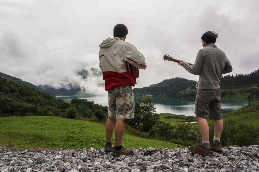 Two people stand on a rocky hill near a lake surrounded by trees and mountains, reminiscent of circumnavigating Mt. Blanc. One holds a guitar, both wear jackets and beanies, looking towards the mist-covered landscape.