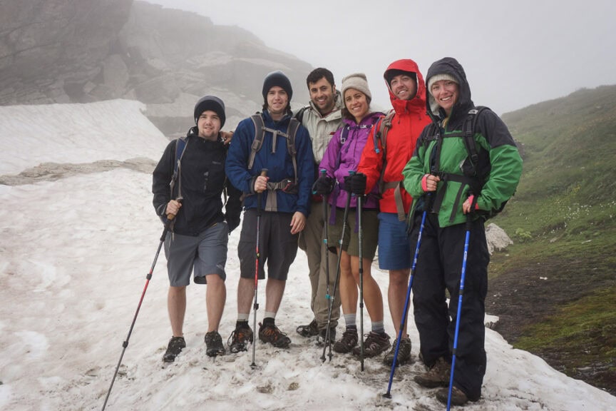 Six hikers in outdoor gear, standing on a snowy mountain trail with trekking poles, are circumnavigating Mt. Blanc. The background is foggy and rocky with a hint of green on the hillside.