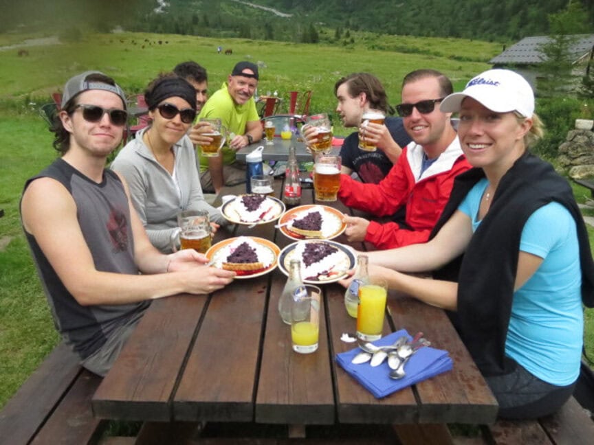 A group of people sit at a wooden picnic table outdoors, holding glasses of beer and enjoying plates of food with Mt. Blanc in the background, recounting their trekking adventures.