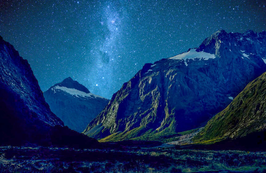 A mountainous landscape at night with a brilliant starry sky and the Milky Way visible above snow-capped peaks, making it one of 5 great places to see.
