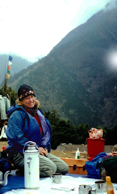 A person with a backpack and hat sits outdoors in front of a mountainous landscape in Nepal, with camping gear and supplies around them, creating moments perfect for a family photobook.