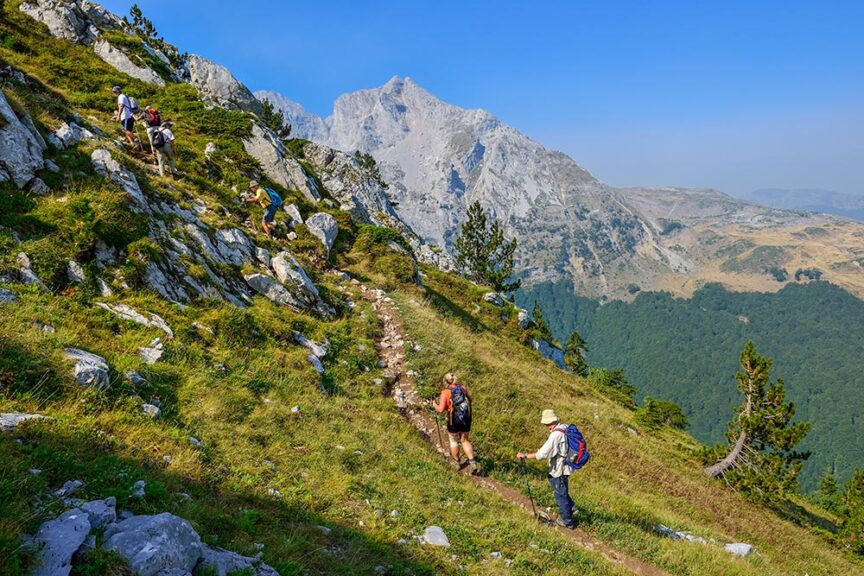 A group of hikers trekking on a steep mountain trail surrounded by rocky terrain and grassy slopes, with a majestic European mountain range in the background under a clear blue sky, blissfully free from crowds.