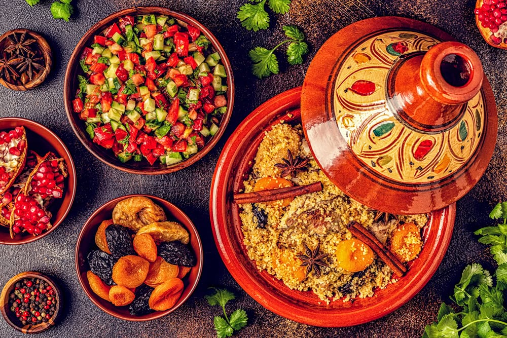 A variety of dishes including a tagine with couscous, a salad with chopped vegetables, dried fruits, and spices are arranged on a table. Green herbs and star anise complete the spread of these delicious recipes.