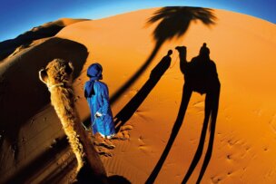 A person in blue stands beside a camel on orange sand dunes, casting long shadows in the setting sun. A shadow of a palm tree is also visible on the sand, making it one of the top places to travel for those seeking a breathtaking sunset experience.