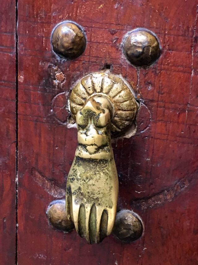 Close-up of a brass door knocker shaped like a hand with a ring, mounted on a weathered wooden door with visible wear and scratches. Exploring Morocco with my best friends, this ancient detail caught our eyes while wandering through the vibrant streets.