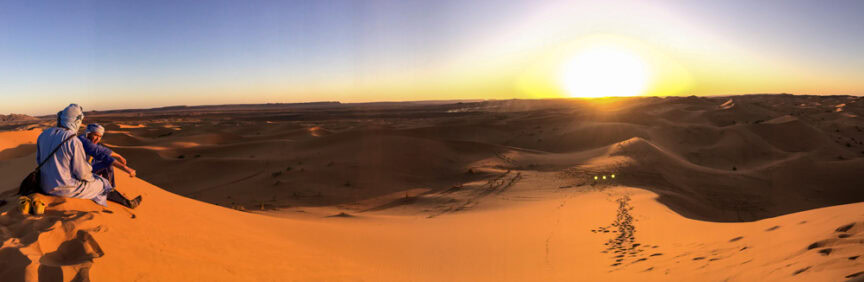 Two best friends, clad in traditional desert clothing, sit atop a sand dune, exploring the vast desert landscape as the sun sets on the horizon, showcasing the best of Morocco.