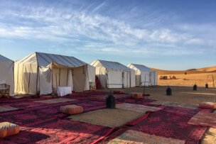 Three white tents are set up in a desert with several red and patterned rugs laid out in front. The sky is clear with a few wispy clouds. Some friends are visible in the distance near the sand dunes, exploring what feels like the enchanting landscapes of Morocco.