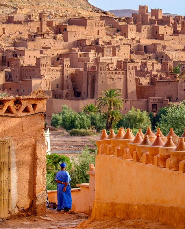 A person in blue traditional clothing stands in front of an ancient clay city with tall, fortified citadels and palm trees, set against a mountainous backdrop.