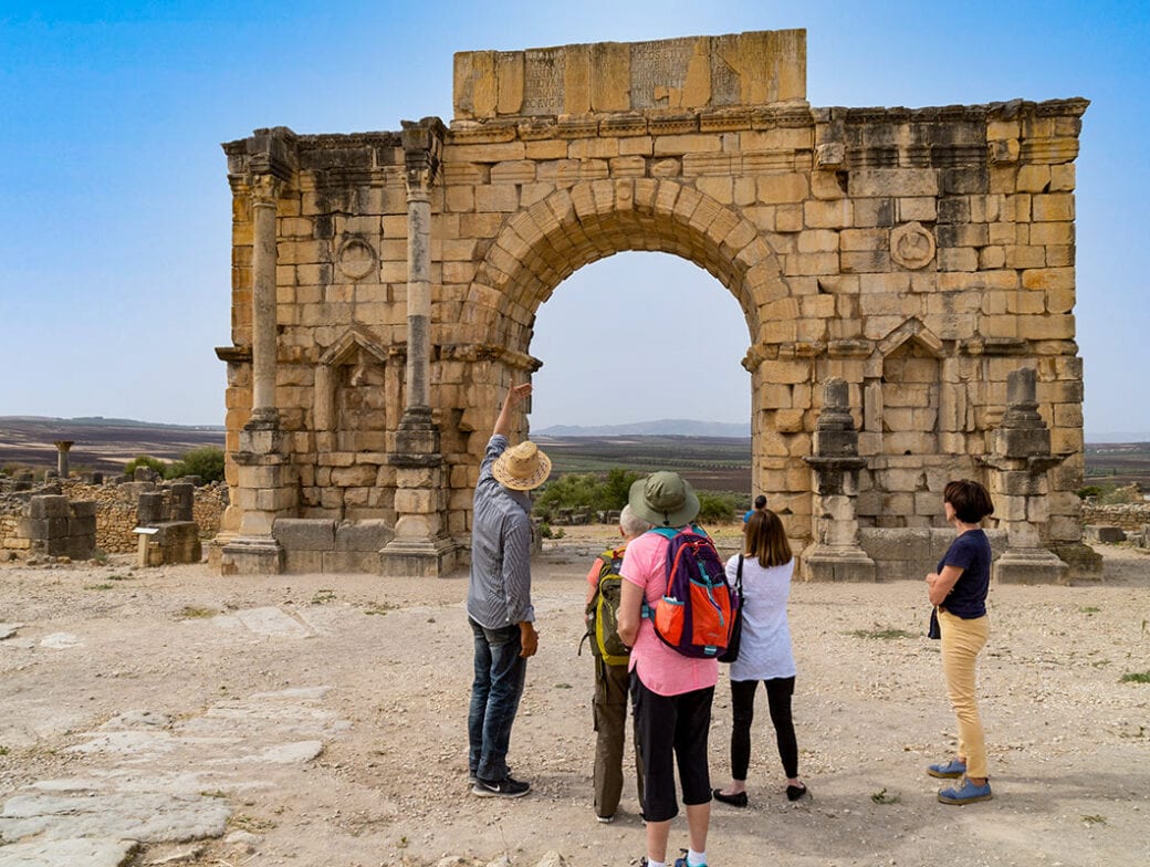 A group of four people stand near a large ancient stone archway in Morocco, with one person pointing towards it. The background features a dry, open landscape under a clear blue sky, capturing the spirit of adventure and hiking through this breathtaking scenery.